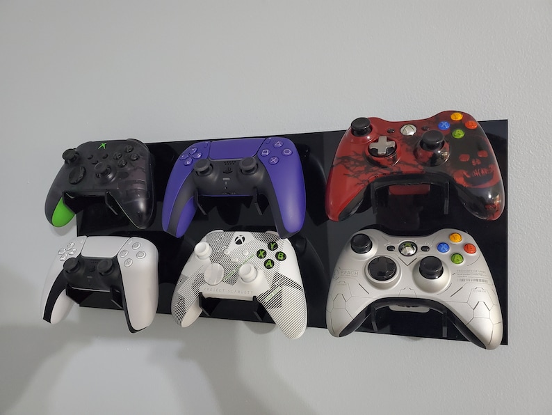 Pode incluir: Seis controladores de videogame diferentes montados em um suporte de parede preto. Os controladores s&atilde;o uma mistura de preto, branco, verde, vermelho e roxo. Os controladores s&atilde;o todos de marcas e modelos diferentes.