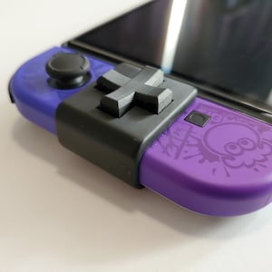 May include: A purple Nintendo Switch console with a black and gray controller attachment. The console has a black screen and a white ink splattered design with a squid-like character.