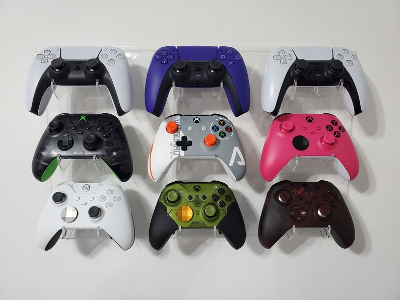 Pode incluir: Uma cole&ccedil;&atilde;o de oito controladores de videogame de diferentes cores, incluindo branco, preto, verde, roxo, rosa e cinza, exibidos em um suporte de parede.