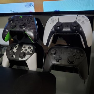 May include: Four different video game controllers are displayed on a black stand. The controllers are black, white, and green. The controllers are arranged in a row from left to right. The controllers are all in a vertical position.