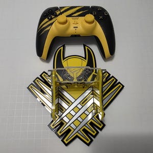 May include: A black and yellow wireless game controller with claw mark graphics positioned above a matching yellow and black wall-mounted display stand featuring a central clear acrylic cradle and a graphic design of three silver metallic claw marks.