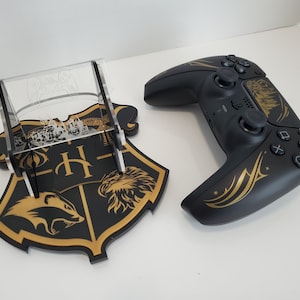 May include: A black and gold video game controller with a Hogwarts house crest design. The controller is on a clear acrylic stand with a Hogwarts house crest base.