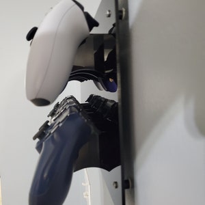 Pode incluir: Um suporte de parede preto com dois controladores de videogame conectados. Um controlador &eacute; branco e o outro &eacute; azul escuro.