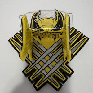 May include: A yellow and black controller stand featuring a graphic of three silver claw marks and the text SNIKT. The stand includes a clear acrylic top piece with an etched controller outline and two yellow vertical supports for holding a device.