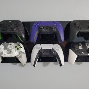 Pode incluir: Seis controladores de videogame diferentes montados em um suporte de parede preto. Os controladores s&atilde;o pretos, brancos, verdes e roxos. Os controladores s&atilde;o de diferentes marcas e modelos.