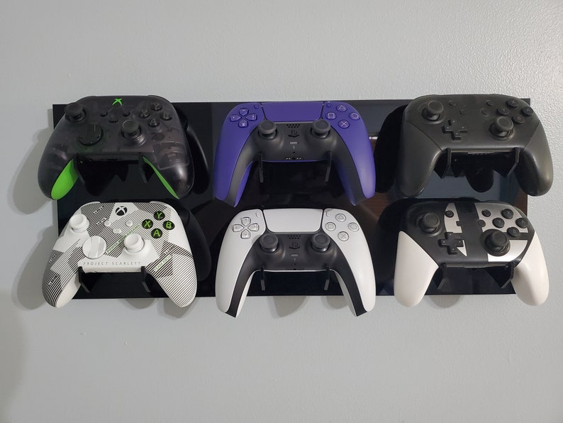 Pode incluir: Seis controladores de videogame diferentes montados em um suporte de parede preto. Os controladores s&atilde;o pretos, brancos, verdes e roxos. Os controladores s&atilde;o de diferentes marcas e modelos.
