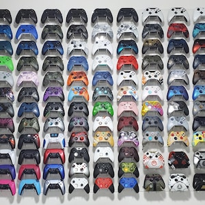 Pode incluir: Uma exposi&ccedil;&atilde;o na parede de mais de 100 controladores de videogame de diferentes cores e padr&otilde;es. Os controladores est&atilde;o dispostos em filas e colunas, com cada fila apresentando uma cor ou padr&atilde;o diferente. Os controladores s&atilde;o todos controladores Xbox, e todos est&atilde;o em perfeitas condi&ccedil;&otilde;es. Os controladores est&atilde;o todos montados em uma parede, e todos est&atilde;o orientados na mesma dire&ccedil;&atilde;o. Os controladores s&atilde;o de v&aacute;rias cores, incluindo preto, branco, vermelho, azul, verde e amarelo. Os controladores s&atilde;o de v&aacute;rios padr&otilde;es, incluindo cores s&oacute;lidas, listras e camuflagem.