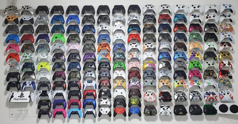Pode incluir: Uma exposi&ccedil;&atilde;o na parede de mais de 100 controladores de videogame de diferentes cores e padr&otilde;es. Os controladores est&atilde;o dispostos em filas e colunas, com cada fila apresentando uma cor ou padr&atilde;o diferente. Os controladores s&atilde;o todos controladores Xbox, e todos est&atilde;o em perfeitas condi&ccedil;&otilde;es. Os controladores est&atilde;o todos montados em uma parede, e todos est&atilde;o orientados na mesma dire&ccedil;&atilde;o. Os controladores s&atilde;o de v&aacute;rias cores, incluindo preto, branco, vermelho, azul, verde e amarelo. Os controladores s&atilde;o de v&aacute;rios padr&otilde;es, incluindo cores s&oacute;lidas, listras e camuflagem.