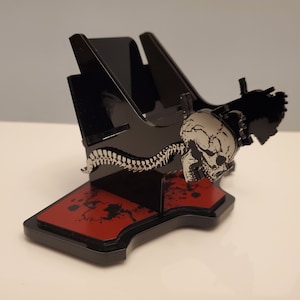May include: Black phone stand with a skull and spine design. The stand has a red base with black splatter paint.