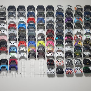 Pode incluir: Uma parede com uma cole&ccedil;&atilde;o de mais de 50 controladores Xbox em v&aacute;rias cores e designs. Os controladores est&atilde;o montados em uma parede branca com suportes de acr&iacute;lico transparente.