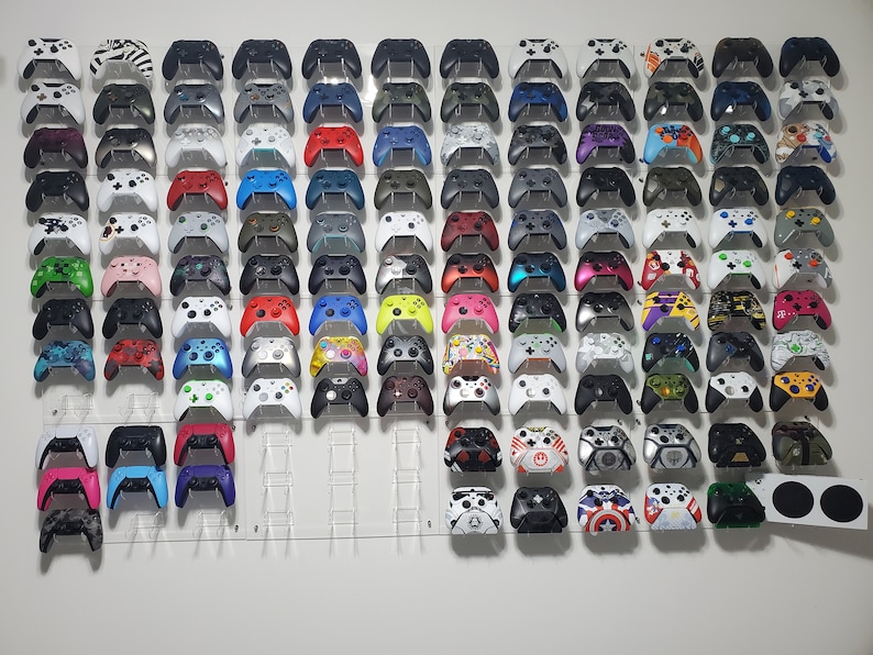 Pode incluir: Uma parede com uma cole&ccedil;&atilde;o de mais de 50 controladores Xbox em v&aacute;rias cores e designs. Os controladores est&atilde;o montados em uma parede branca com suportes de acr&iacute;lico transparente.
