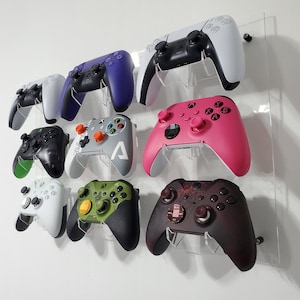 Pode incluir: Um expositor de parede com oito controladores de videogame de diferentes cores. Os controladores s&atilde;o montados em um painel de acr&iacute;lico transparente. Os controladores s&atilde;o de diferentes cores, incluindo preto, branco, verde, rosa, roxo e cinza.