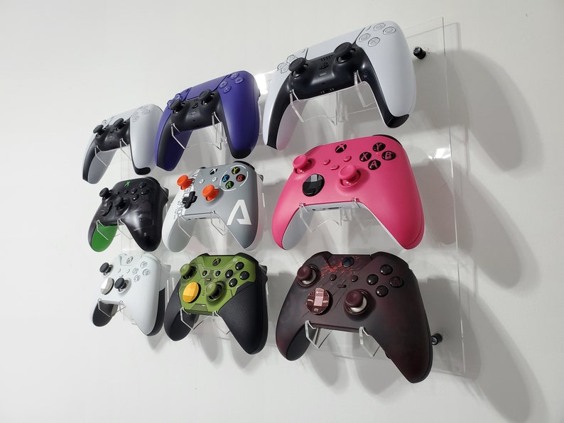 Pode incluir: Um expositor de parede com oito controladores de videogame de diferentes cores. Os controladores s&atilde;o montados em um painel de acr&iacute;lico transparente. Os controladores s&atilde;o de diferentes cores, incluindo preto, branco, verde, rosa, roxo e cinza.