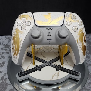 May include: A white and gray PlayStation 5 controller with gold brushstroke accents, displayed on a stand featuring crossed katana swords. The controller has a gold design on the top and sides.