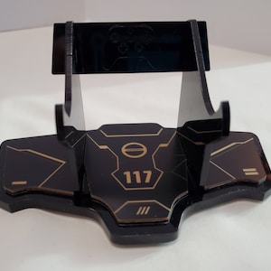 May include: Black acrylic stand with a gold design featuring the number 117. The stand is designed to hold a video game controller.