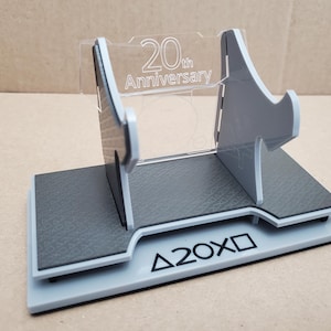 May include: A gray and black display stand with the text "20th Anniversary" on a clear plastic panel. The base has the text "Δ20XD". The stand is designed to hold a gaming console or related item.