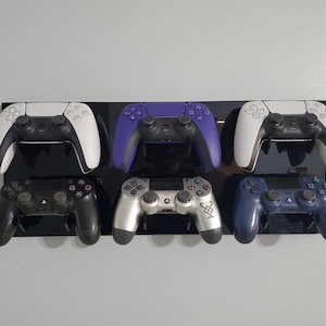 Pode incluir: Seis controladores de videogame de diferentes cores est&atilde;o montados em um suporte de parede preto. Os controladores s&atilde;o branco, roxo, preto, prata, azul e branco.