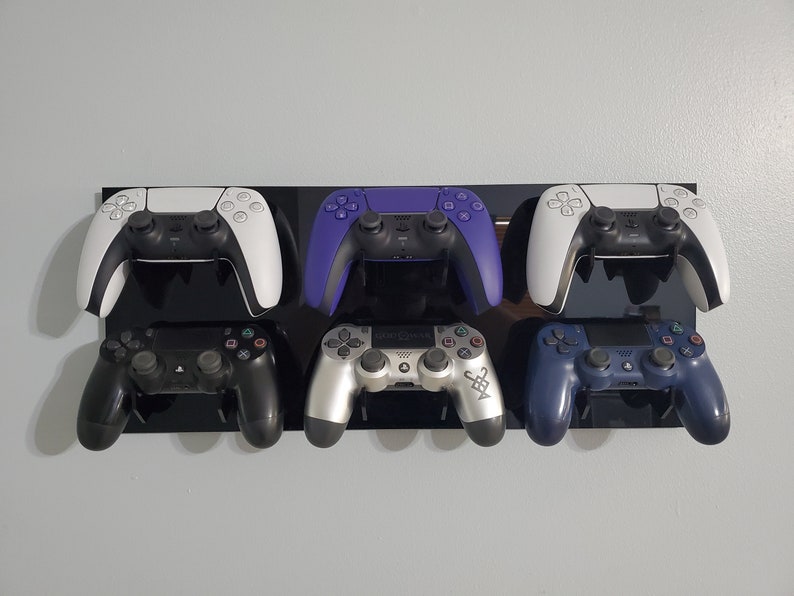 Pode incluir: Seis controladores de videogame de diferentes cores est&atilde;o montados em um suporte de parede preto. Os controladores s&atilde;o branco, roxo, preto, prata, azul e branco.