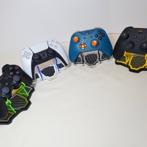 May include: Four different video game controllers on stands. The controllers are black, white, blue, and black. The stands are black and green.