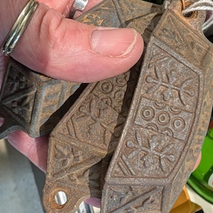 May include: A collection of antique, cast iron decorative pieces with intricate floral and geometric patterns. The pieces are a rusty brown color and have a loop at the end for hanging. The design suggests they may be decorative hardware or architectural elements.