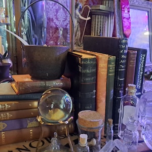 May include: A collection of vintage books, antique bottles, and a cauldron on a wooden shelf. The books have titles like "The Book of Black Magic" and "The Egyptian Book of the Dead".
