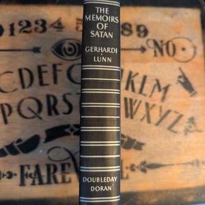 May include: A hardcover book titled "The Memoirs of Satan" by Gerhardi Lunn. The spine is dark gray with white horizontal stripes and text. The publisher is Doubleday Doran. The book is set against a wooden background.