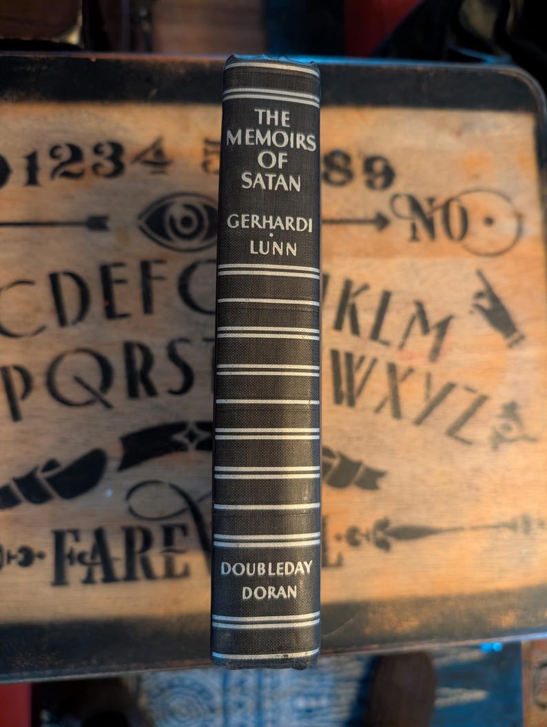 May include: A hardcover book titled "The Memoirs of Satan" by Gerhardi Lunn. The spine is dark gray with white horizontal stripes and text. The publisher is Doubleday Doran. The book is set against a wooden background.