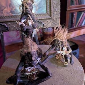 May include: Three dark wooden figurines with straw hair and accents. One figure stands holding drumsticks and a drum. The other two figures are in a squatting position. The figurines are on a round, light-colored surface.