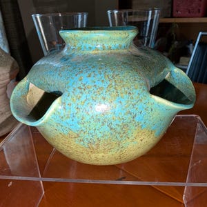 May include: A turquoise and yellow speckled ceramic vase with three openings. The vase has a rounded shape with a flared rim and is displayed on a clear acrylic stand. The vase has a speckled texture.