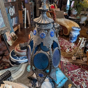 May include: An ornate, antique metal lantern with blue glass panels and decorative filigree. The lantern has a silver finish and is adorned with blue accents. The design is intricate and detailed, suggesting a vintage or gothic style. The lantern is a decorative item.