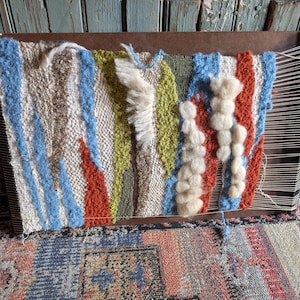 May include: A colorful woven textile art piece in progress, mounted on a wooden frame. The artwork features vertical stripes in blue, orange, green, and white, with textured, raised elements. The weaving is still attached to the loom.