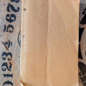 May include: A rectangular, light tan paper booklet with a smooth, matte texture. The cover features two small, dark brown circular punch holes aligned vertically along the left edge. The paper shows signs of wear, including a creased corner.
