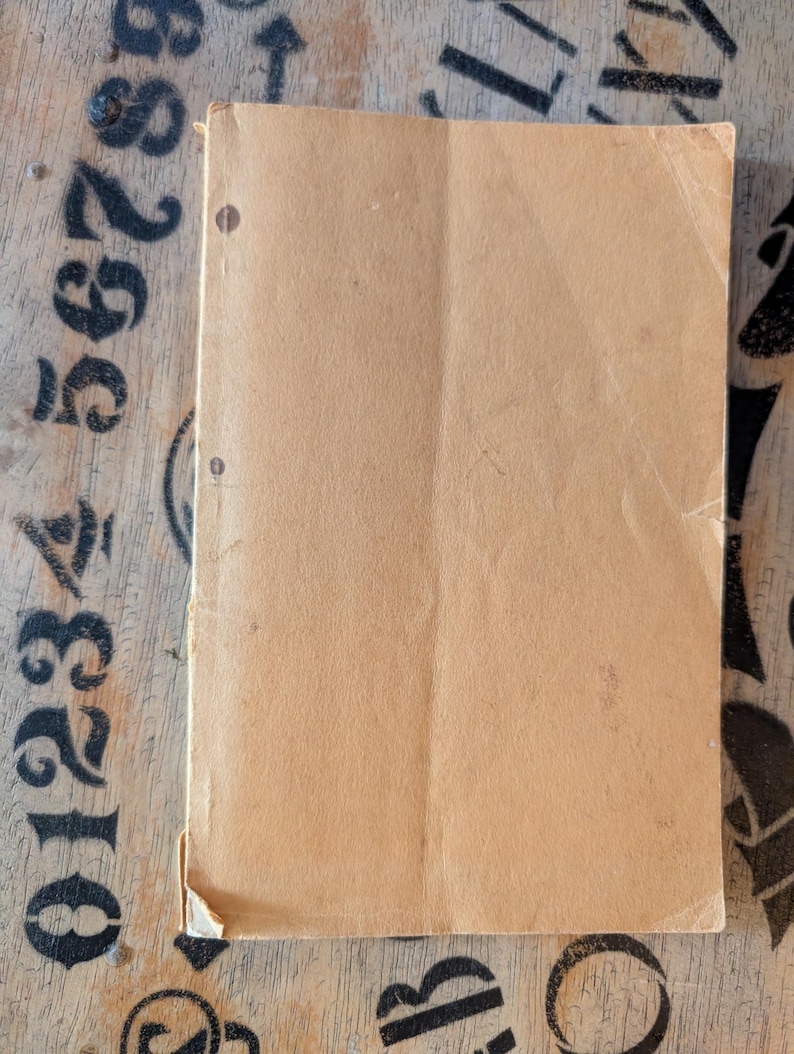 May include: A rectangular, light tan paper booklet with a smooth, matte texture. The cover features two small, dark brown circular punch holes aligned vertically along the left edge. The paper shows signs of wear, including a creased corner.
