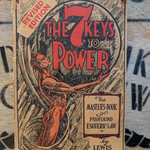 May include: A vintage book titled The 7 Keys to Power by Lewis de Claremont. The cover features a red illustration of a figure emerging from a vessel, with the text Revised Edition, The Master's Book of Profound Esoteric Law, and Price $2.00.