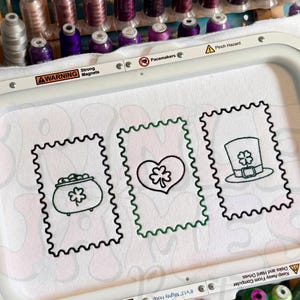 May include: An embroidery hoop holding white fabric with three St. Patrick's Day themed designs: a pot of gold, a heart with a shamrock, and a leprechaun hat. The hoop is surrounded by spools of thread in various colors.