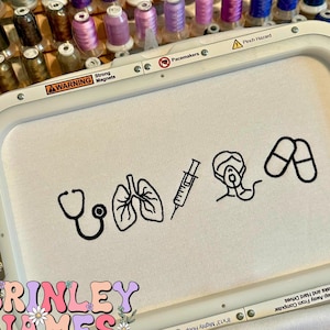 Puede incluir: Tela blanca con símbolos médicos bordados en negro, incluyendo un estetoscopio, pulmones, una jeringa, una mascarilla y pastillas. La tela está sujeta en un bastidor de bordado blanco. La imagen también muestra carretes de hilo de varios colores.