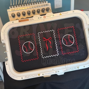 May include: An embroidery machine with a black fabric featuring three postage stamp designs. Two stamps have baseballs, and one has a red bow. The word "Boutique" is visible on the fabric.