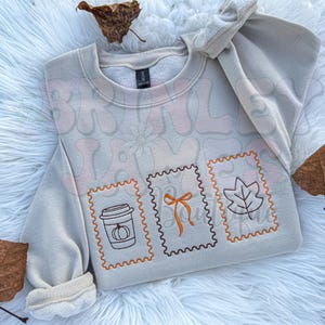 May include: A beige sweatshirt with a fall-themed design. The design includes three postage stamp-like images with a coffee cup, a bow, and a leaf. The sweatshirt has a crew neck and long sleeves. The background is a white, fluffy surface.