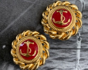 May include: A pair of gold-tone earrings with red enamel anchors in the center. The anchors are surrounded by a rope-like design.