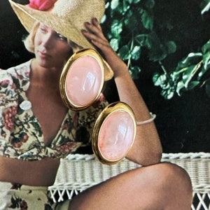 May include: A pair of gold tone earrings with pink oval shaped stones. The earrings are shown on a model wearing a floral dress.