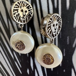 May include: A pair of silver tone drop earrings with a lion head design and a white pearl-like drop. The lion head is surrounded by a circle of raised lines.
