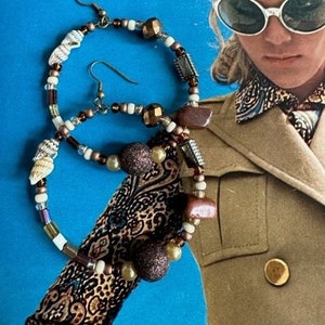May include: A pair of hoop earrings with a variety of beads in shades of brown, gold, and white. The earrings also feature small seashells and glittery accents. The earrings are displayed on a blue background.