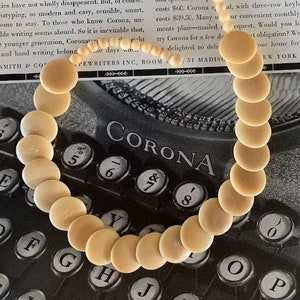 May include: A vintage ivory-colored necklace with large, round beads. The necklace is laid out on a black and white typewriter with the word "CORONA" visible on the keys.