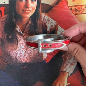 May include: A red and silver bangle bracelet with a buckle detail. The bracelet is made of leather and metal.
