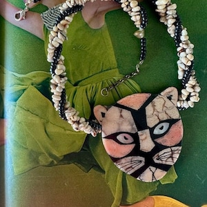 May include: A white and black beaded necklace with a large, pink and black ceramic pendant shaped like a cat's face.