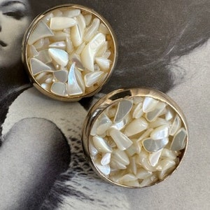 May include: Two round, vintage earrings with gold-tone metal rims. Each earring is filled with iridescent, irregularly shaped pieces, creating a mosaic effect. The earrings are set against a vintage black and white background.