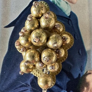 May include: A gold-toned metal brooch with a cluster of round, textured beads. The beads are arranged in a geometric pattern and have a slightly raised, textured surface.