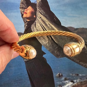May include: A gold-tone cuff bracelet featuring a twisted cable design and two polished gold-tone end caps, each set with a smooth, round white pearl. The bracelet has an open-ended structure, allowing for easy placement on the wrist.