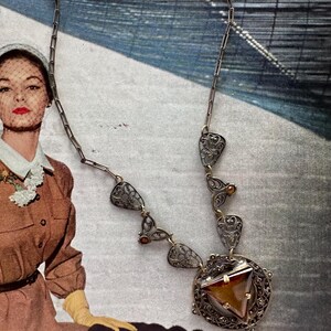 May include: A vintage silver-toned necklace with a triangular amber-colored pendant. The necklace features ornate filigree details and a delicate chain. The pendant is set within a circular frame, showcasing a retro aesthetic.