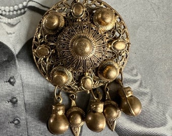 May include: A large, round, gold-toned brooch with intricate filigree detailing and multiple dangling round ornaments. The brooch has a central circular design with a raised, textured center.
